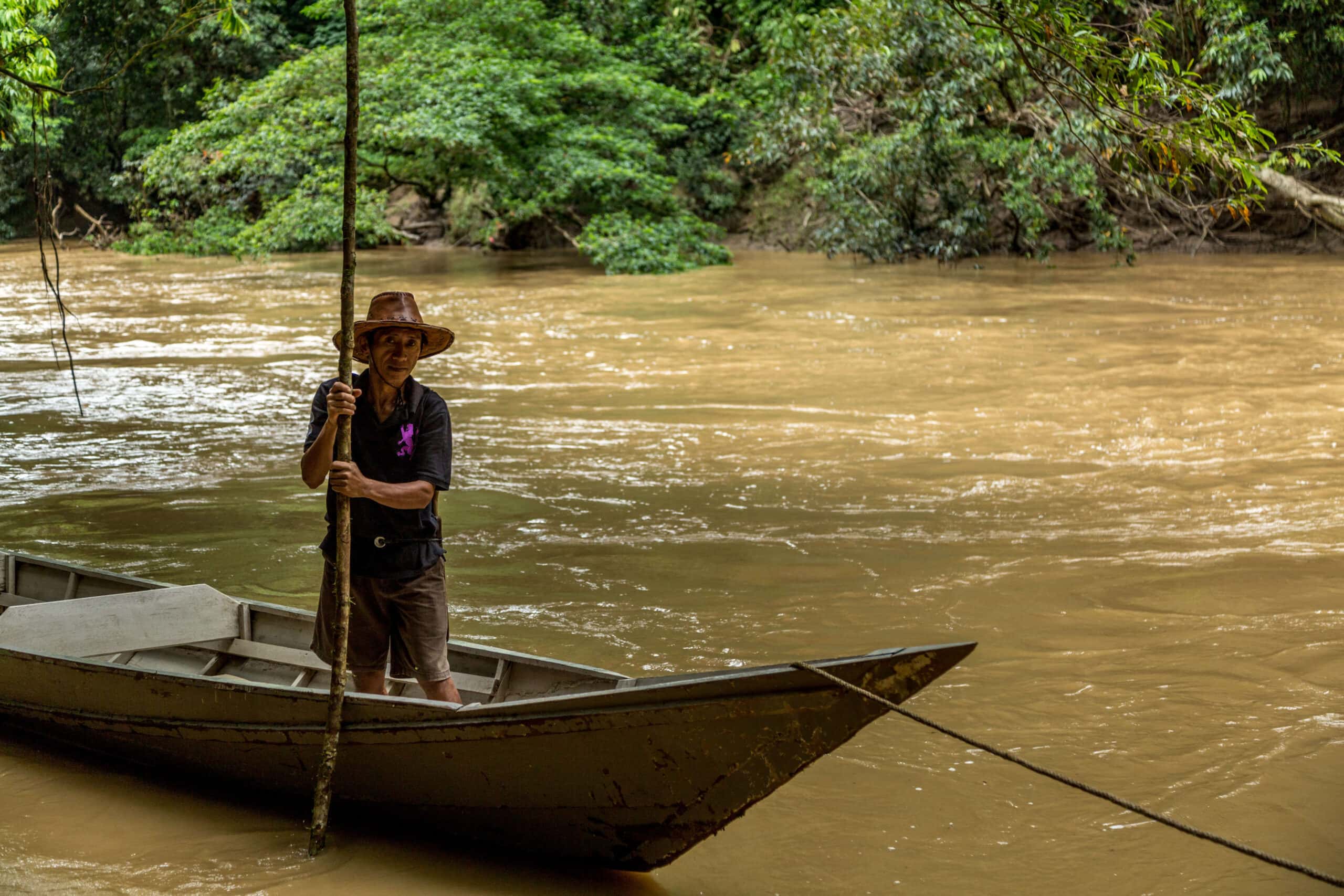 Man standing in wooden boat in the Kinabatangan River Man standing in wooden boat in the Kinabatangan River with jungle backdrop in Borneo
